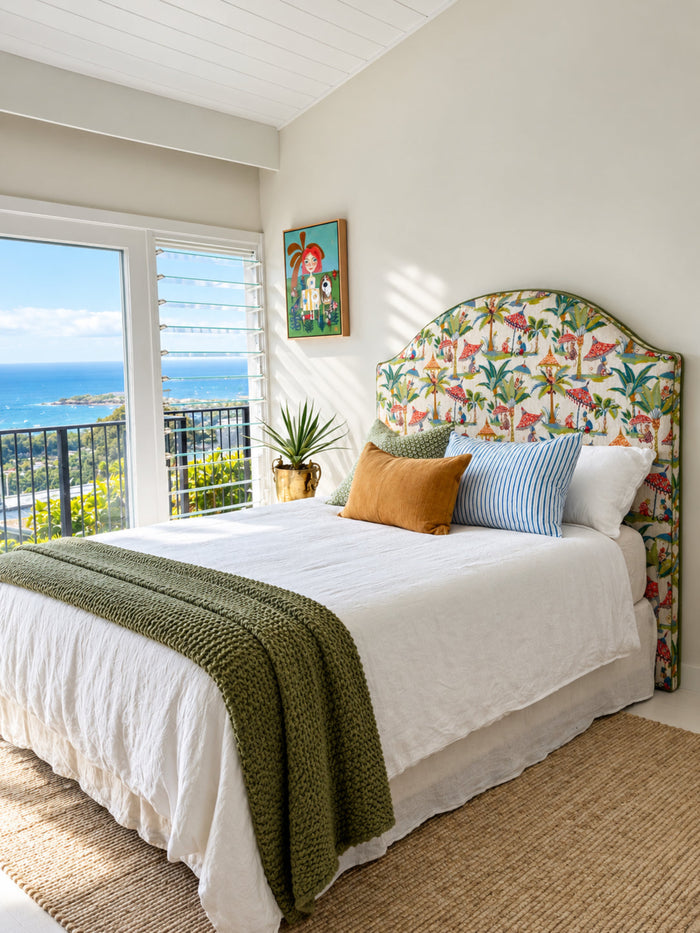 A bed styled in a bright coastal bedroom, featuring a tropical bird-print upholstered headboard, white bedding, a green knit throw, and colorful accent pillows. A large window offers a view of the blue ocean.