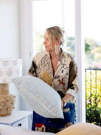 Woman holding a decorative pillow in a bright room with large windows.