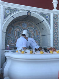 Two individuals in white lab coats standing behind a decorative fountain with fruit and a person observing from the side.