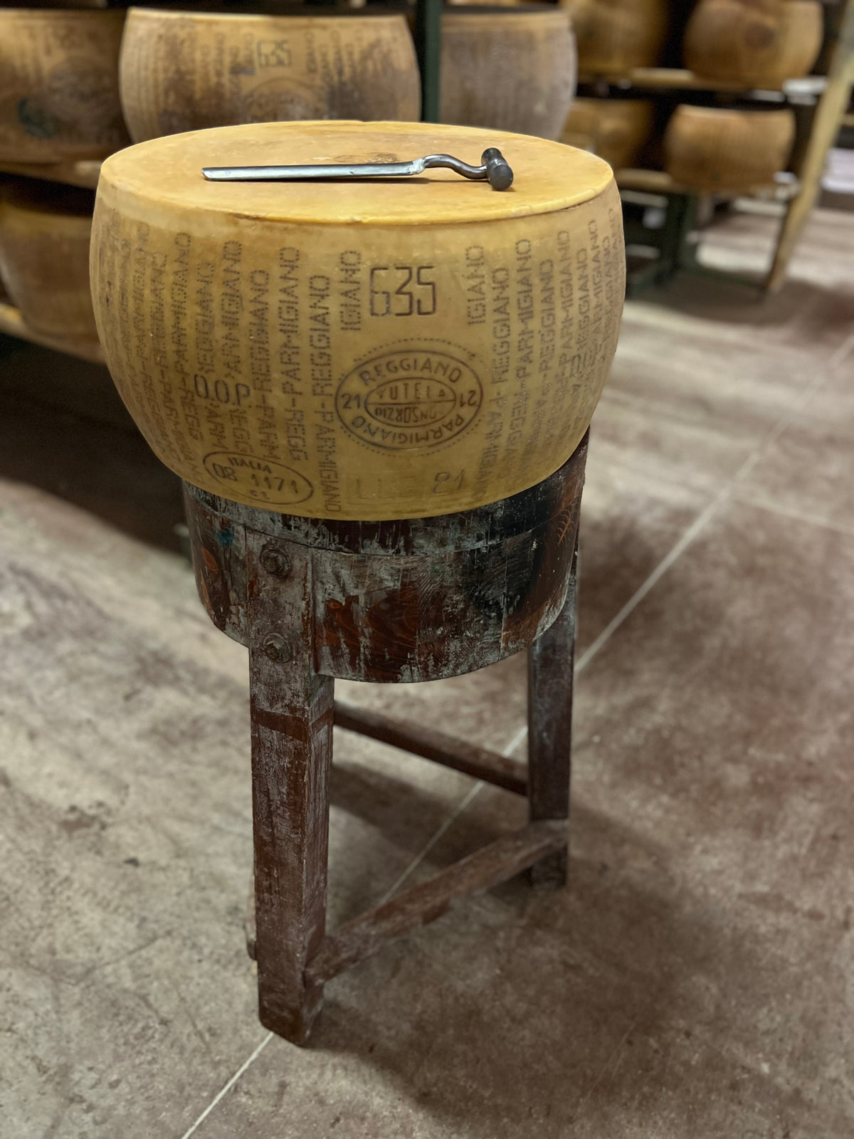 Wooden stool made from a Parmigiano-Reggiano cheese wheel with a metal pin, in a warehouse setting.