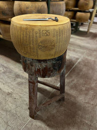 Wooden stool made from a Parmigiano-Reggiano cheese wheel with a metal pin, in a warehouse setting.