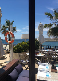 Outdoor seating area with umbrellas, tables, and chairs near a waterfront with palm trees.