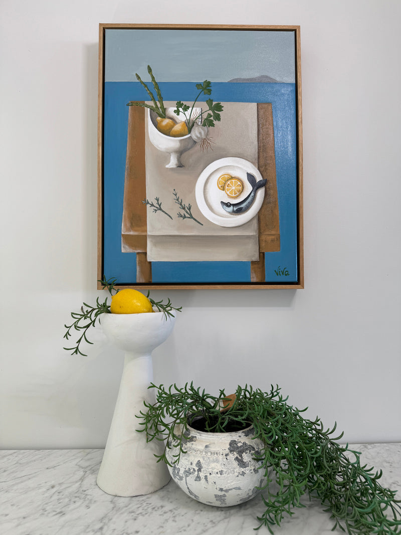 Framed artwork with a tea set and lemon next to a white teapot with greenery on a light gray wall.
