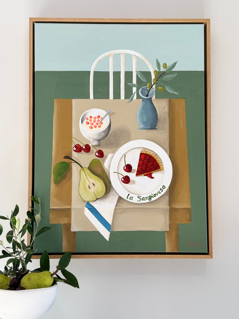 Framed artwork of a table setting with food and a chair on a light green background