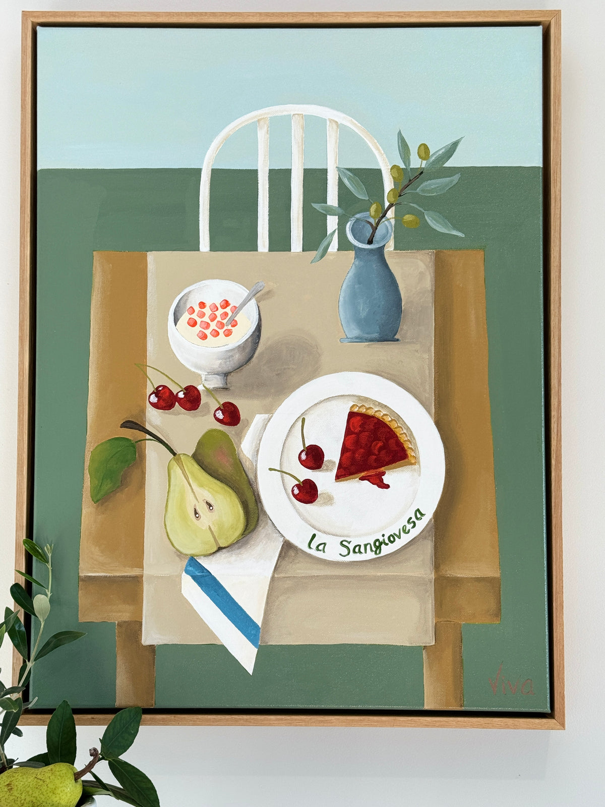 Framed artwork of a table setting with food and a chair on a green background