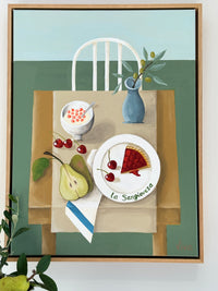 Framed artwork of a table setting with food and a chair on a green background