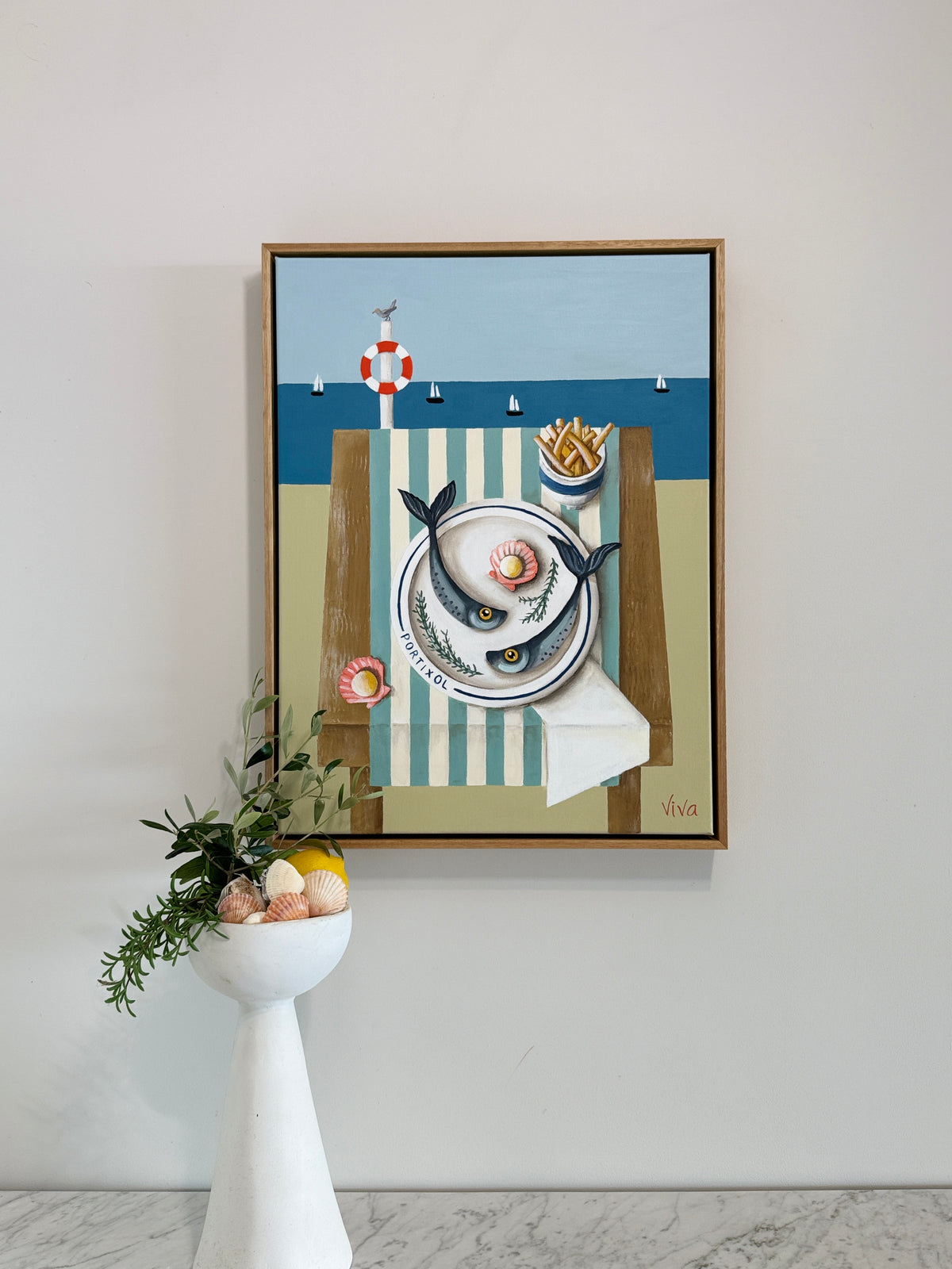 Framed artwork of a cat with a plate of food on a chair against a beach scene background, displayed on a wall.