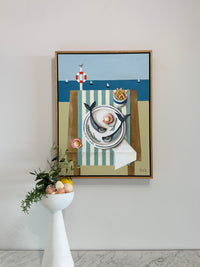 Framed artwork of a cat with a plate of food on a chair against a beach scene background, displayed on a wall.