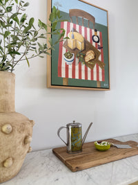 Framed artwork on a wall above a marble surface with a teapot and cutting board.