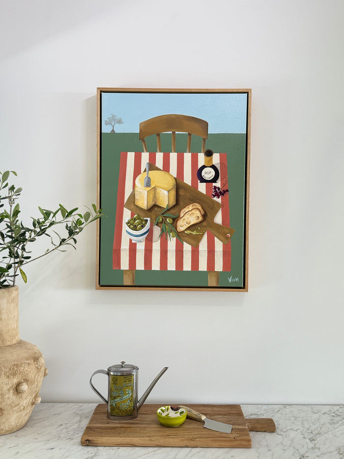 Framed artwork of a table setting with food on a striped tablecloth, placed on a white wall above a wooden cutting board with a teapot and knife.