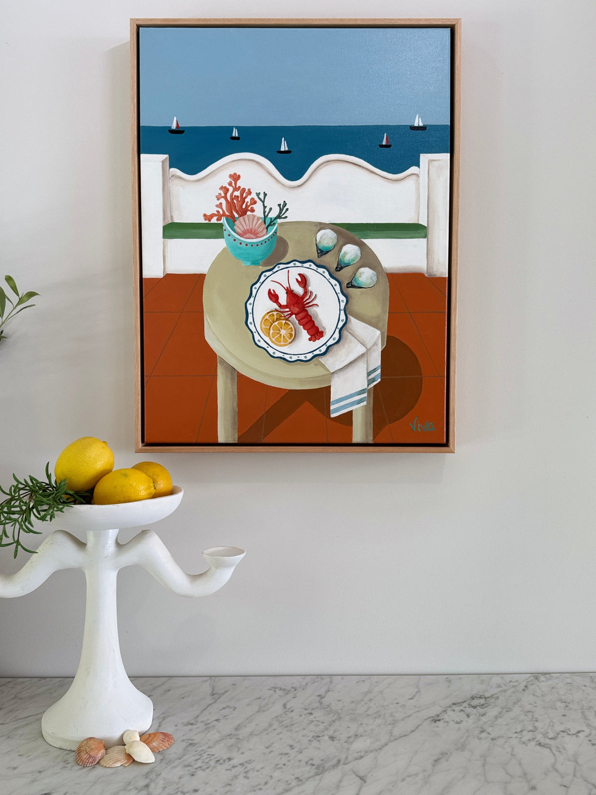 Framed artwork of a table with a lobster plate and flowers, placed on a wall above a white pedestal with lemons.