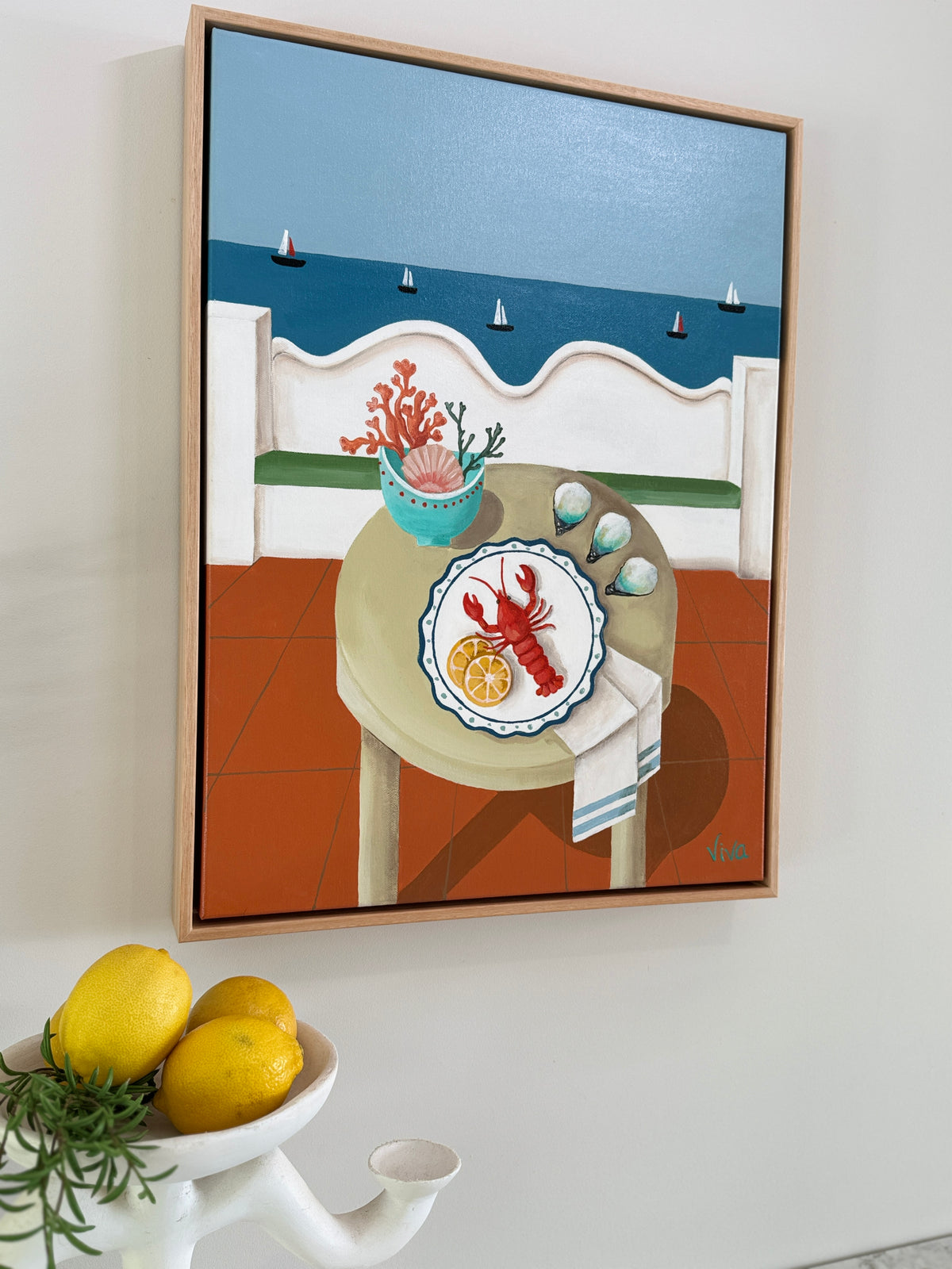 Framed artwork of a table with a lobster plate on a white wall, with lemons and a teapot in the foreground.