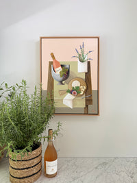 Framed artwork on a wall with a bottle of wine and a plant in the foreground.