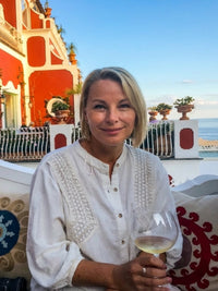 Woman holding a glass of wine on a balcony with ocean view and decorative cushions.