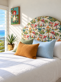 A styled bed in a bright bedroom featuring a large arched headboard upholstered in a colorful tropical print with palm trees, beach umbrellas, and figures. The bed is made with white bedding, a mustard yellow pillow, a green textured pillow, and a blue-and-white striped pillow.