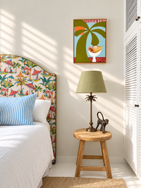 A styled bedroom corner with a bed featuring a colorful tropical flamingo-print headboard, a wooden stool with a green lamp and small monkey figurine, and a framed art print on the wall above.