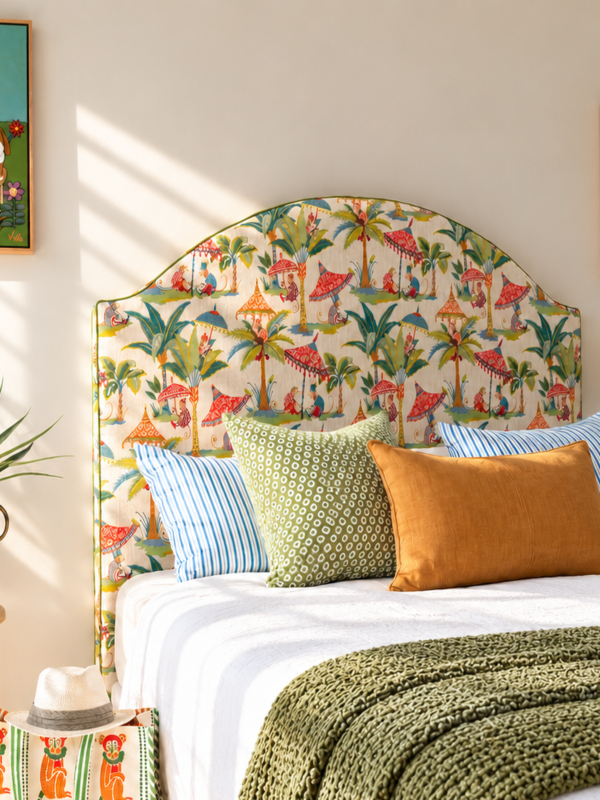 A tropical-themed bedroom featuring a bed with a colorful palm tree and flamingo print upholstered headboard, styled with blue and green pillows, an orange accent pillow, a green knit throw blanket, and two framed art prints on the beige walls.