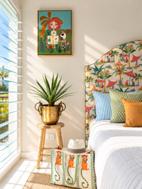 A styled bedroom corner with a bed featuring a colorful tropical flamingo-print upholstered headboard, matching decorative pillows, a small wooden stool holding a potted plant, and a patterned bag on the floor. A framed art print hangs on the wall above.