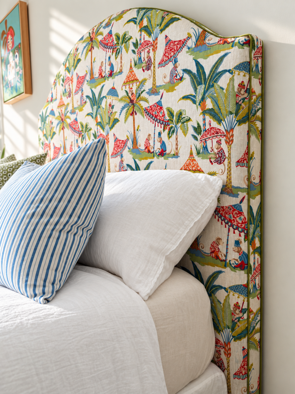 A close-up of a bed with a tall upholstered headboard covered in a colorful tropical print featuring palm trees, birds, and figures on a cream background. White bedding and pillows are visible, along with a blue-and-white striped accent pillow.