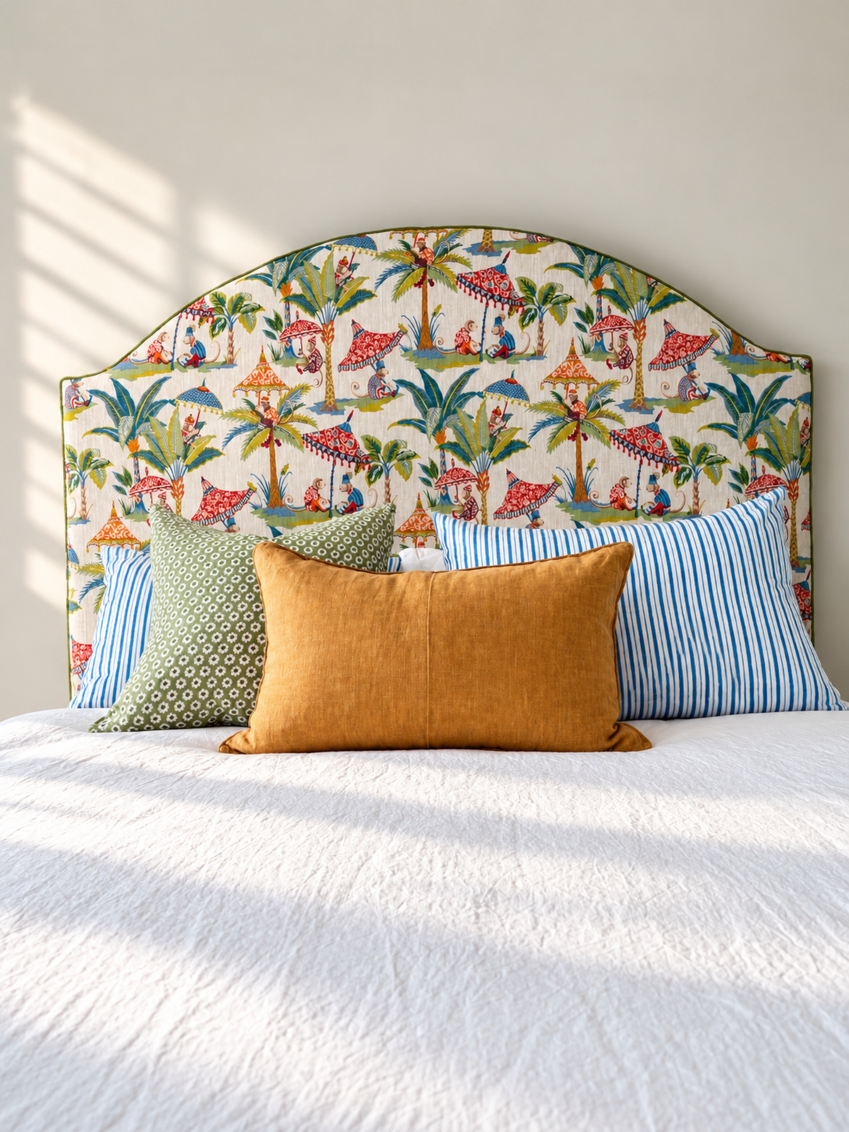A bed styled with a colorful tropical-themed upholstered headboard, a mustard yellow linen pillow, a green patterned pillow, and blue striped pillows against a neutral wall with soft window light.