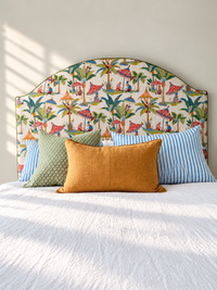 A bed styled with a colorful tropical-themed upholstered headboard, a mustard yellow linen pillow, a green patterned pillow, and blue striped pillows against a neutral wall with soft window light.