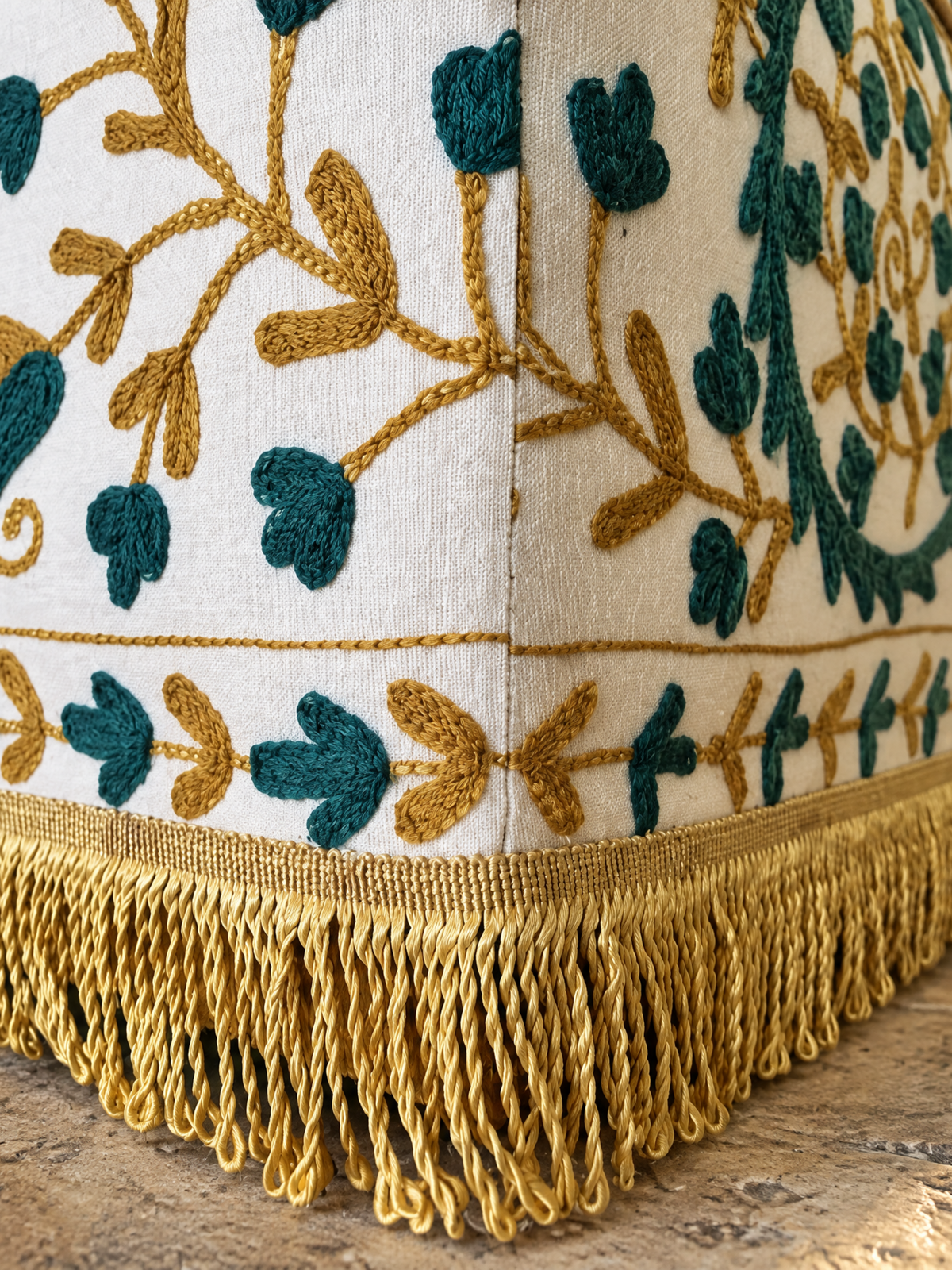 Close-up of a decorative upholstered cushion corner with a cream fabric base, teal and mustard embroidered floral vine pattern, and a long gold fringe along the bottom edge.