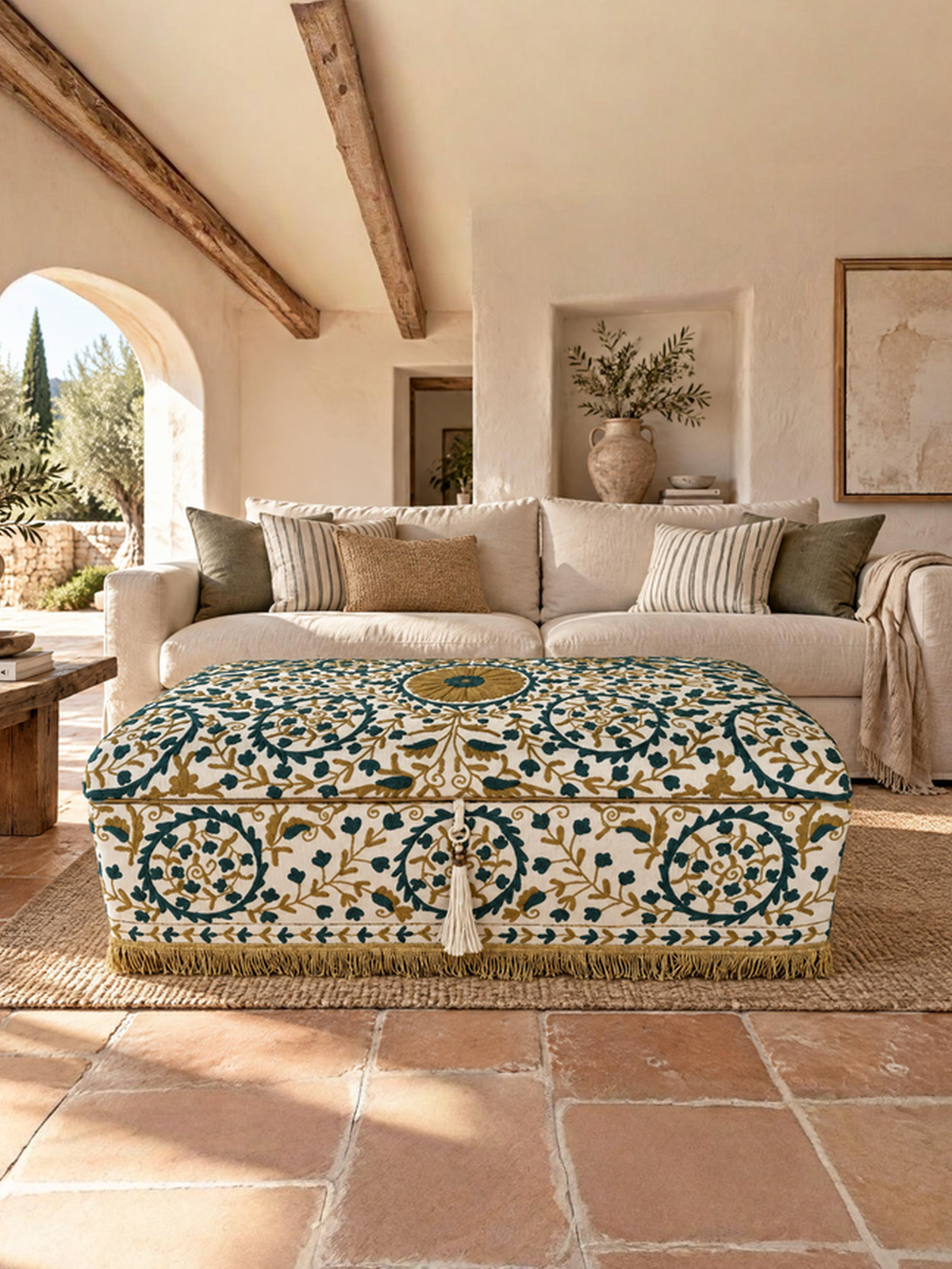 A rectangular ottoman with a detailed blue, green, and gold floral pattern on a cream background, styled in a rustic, sunlit living room with terracotta tile floors and a beige sofa.