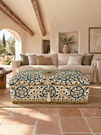 A rectangular ottoman with a detailed blue, green, and gold floral pattern on a cream background, styled in a rustic, sunlit living room with terracotta tile floors and a beige sofa.