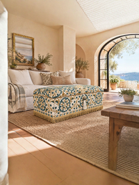 A styled living room interior featuring a decorative storage ottoman with a blue, green, and cream floral pattern and gold fringe, positioned on a large woven jute rug in front of a cream-colored sofa.