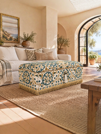 A styled living room interior featuring a decorative storage ottoman with a blue, green, and cream floral pattern and gold fringe, positioned on a large woven jute rug in front of a cream-colored sofa.