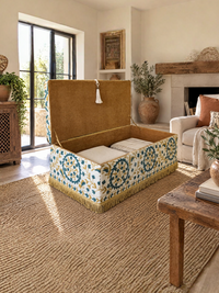 A decorative storage ottoman with a tan suede-like top and a blue, gold, and white patterned base, styled in a bright, modern living room.