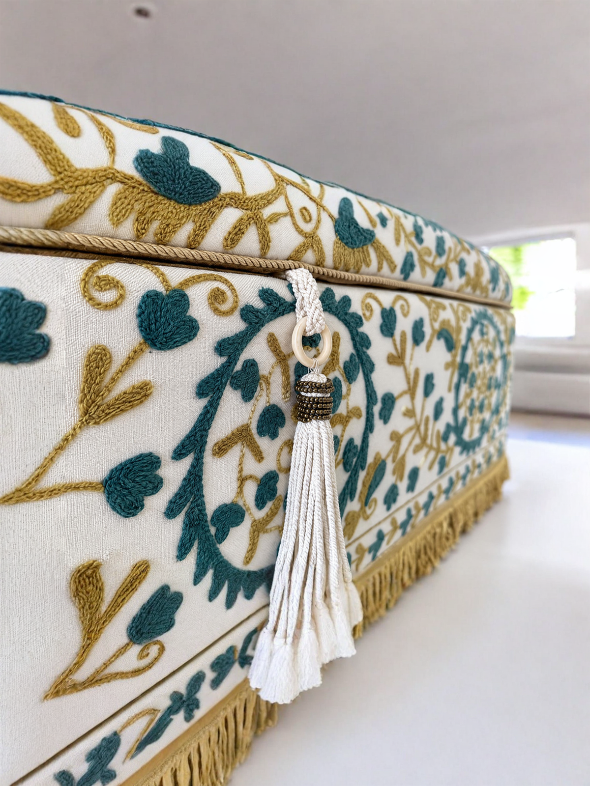 Close-up of an upholstered storage box or bench corner with cream fabric, teal floral embroidery, gold vine embroidery, and a white tassel hanging from a rope handle.